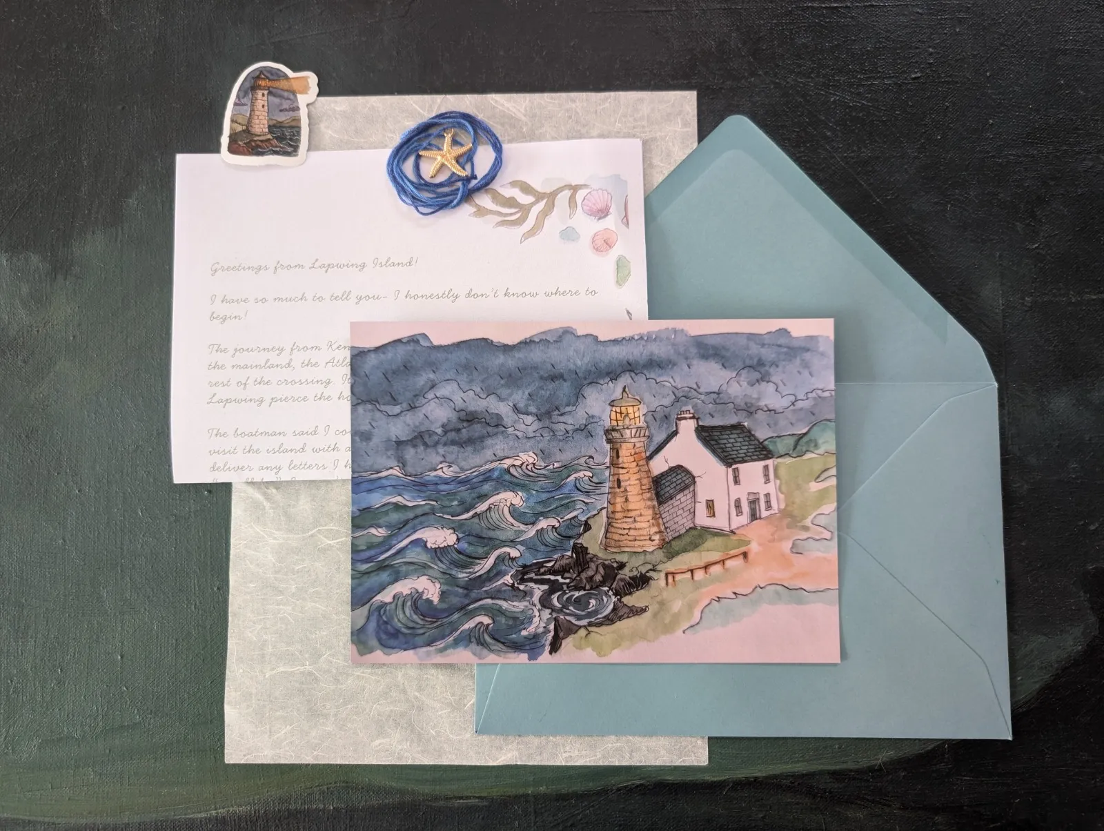 An open letter from Lapwing Island beside a watercolour postcard of a lighthouse, a teal envelope, a starfish charm and a lighthouse sticker.